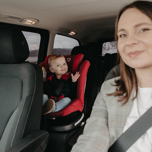 Fröhliches Kleinkind winkt aus einem roten Kindersitz, während eine Frau im Autofahrersitz lächelt.