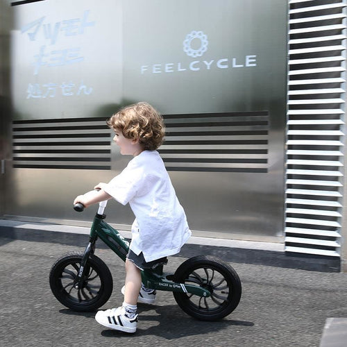 Kind in Bewegung auf einem grünen Laufrad, vor einer urbanen Kulisse mit Schriftzug 'FEELCYCLE'.