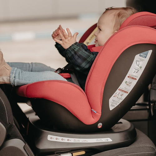 Lächelndes Kleinkind klatscht in einem roten Kindersitz, Sicherheitsgurt ist angelegt, grauer Autositz im Hintergrund.