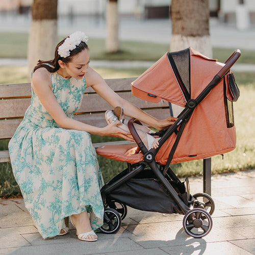 Eine Frau im blumigen Sommerkleid sitzt auf einer Bank und beugt sich über einen korallenfarbenen Reisebuggy.