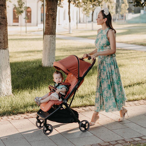 Eine Frau in einem sommerlichen Kleid schiebt einen orangefarbenen Reisebuggy, in dem ein Kleinkind sitzt, auf einem sonnigen Parkweg.