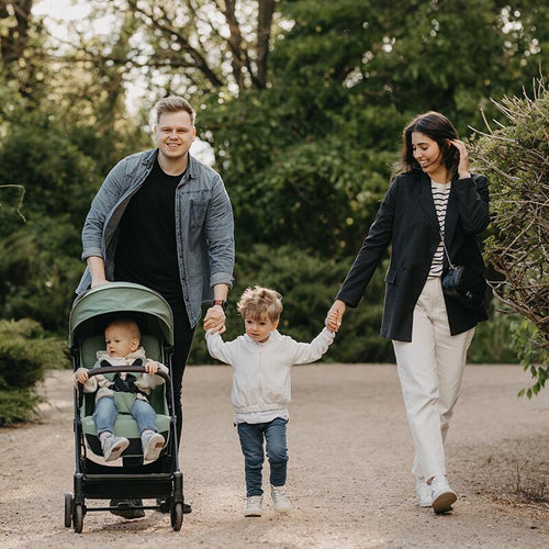 Familie spaziert glücklich, ein Kind im Reisebuggy, das andere hält Elternhand.