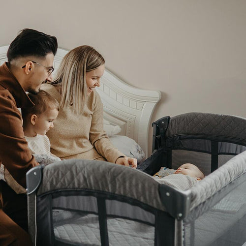 Eine Familie mit einem Kleinkind schaut gemeinsam auf ein Baby, das im Coccolle Insieme Reisebett liegt, Farbe Greystone.