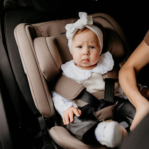 Unzufrieden schauendes Baby im taupefarbenen Kindersitz im Auto, neben ihm eine weibliche Hand.