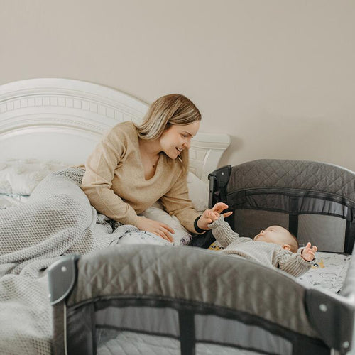 Eine Mutter interagiert mit ihrem auf dem Rücken liegenden Baby, das sich in einem Coccolle Insieme Reisebett befindet.