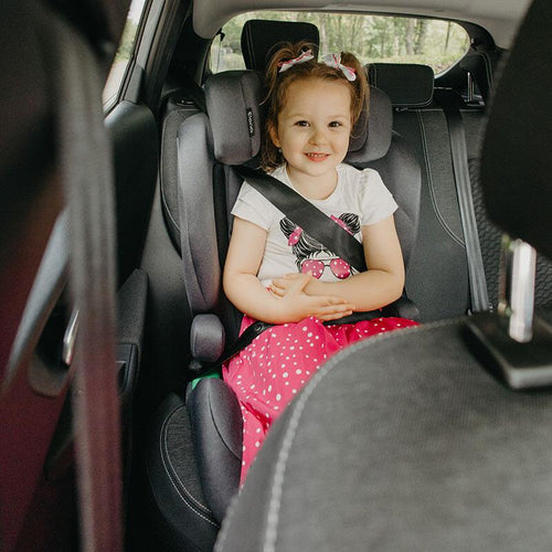 Fröhliches Kind in einem grausteinfarbigen Kindersitz lächelt breit, trägt ein verspieltes Outfit mit Katzenmotiv, im Auto sitzend.