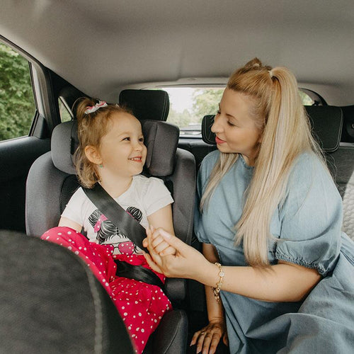 Glückliches junges Mädchen mit Schleifen in den Zöpfen interagiert mit einer Frau, die neben ihr im Auto sitzt, beide lächeln, das Kind sitzt in einem grausteinfarbigen Kindersitz.