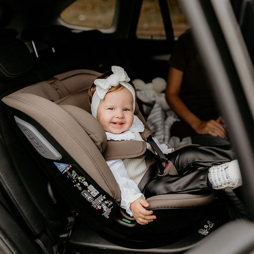 Lächelndes Baby mit weißem Haarband sitzt in einem taupefarbenen Kindersitz im Auto.