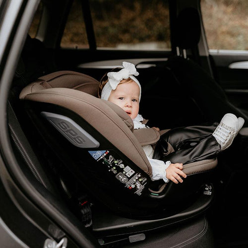 Nachdenklich schauendes Baby in taupefarbenem Kindersitz, Tür des Autos offen, innen sichtbar.