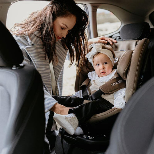 Eine Frau beugt sich in ein Auto, um ein Kleinkind mit einer weißen Schleife im Haar in einem braunen Coccolle-Kindersitz zu sichern.