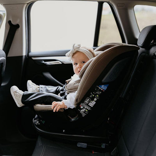 Baby blickt aus einem taupefarbenen Kindersitz im Auto zur Seite, offenes Fenster zeigt den Himmel.