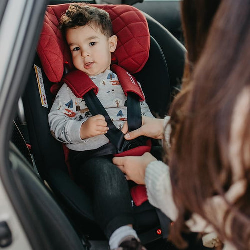 Kleinkind lächelt, angeschnallt in einem roten Kindersitz, Mutter schnallt Sicherheitsgurt im Auto.