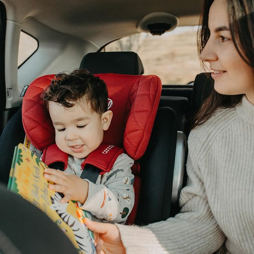 Fröhliches Kind im roten Kindersitz hält ein buntes Buch, Mutter blickt es an, helle Innenausstattung des Autos.