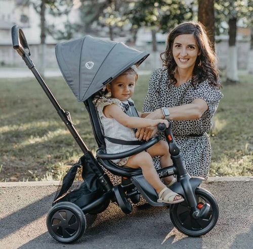 Ein Kleinkind sitzt in einem schwarzen Dreirad-Kinderwagen, lächelnd neben einer Frau mit einem gepunkteten Kleid in einem schattigen Park.