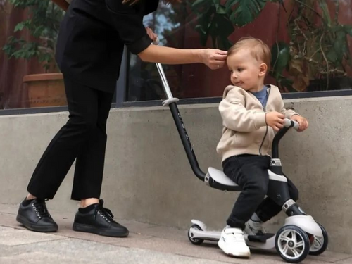 Ein Kleinkind fährt auf einem grauen Scooter, während eine Person in schwarzer Kleidung sich vorbeugt, um es zu stützen.